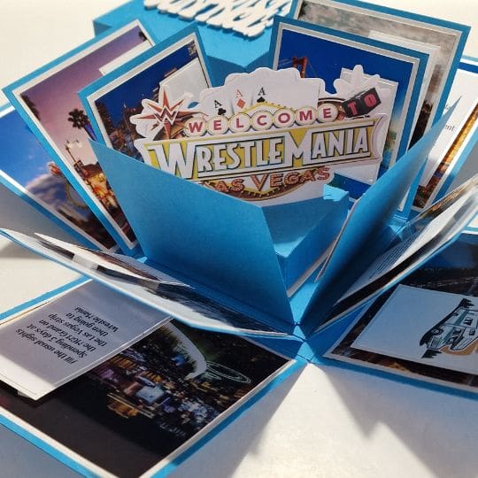 The image shows a pop-up card with a 'Welcome to WrestleMania Las Vegas' theme. The card is blue and has multiple panels that open up to reveal various images and text. The central part of the card features a prominent 'WrestleMania' sign, while the surrounding panels display different pictures, likely related to Las Vegas and WrestleMania events. One of the panels contains text that reads: 'This is your ticket to WrestleMania 38 in Las Vegas, NV on April 3rd, 2022. Enjoy the show